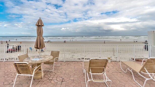 Una terraza con mesa y sillas, con vista a una playa donde hay gente y un cielo nublado.