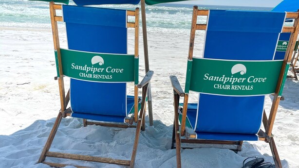 Deux chaises de plage, avec un parasol vert et bleu portant l’inscription « Sandpiper Cove Chair Rentals », sont installées sur une plage de sable, avec l’océan en arrière-plan.