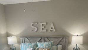 Une chambre à coucher avec un lit, deux tables de chevet, une lampe fixée au mur et le mot décoratif « MER » au-dessus du lit.