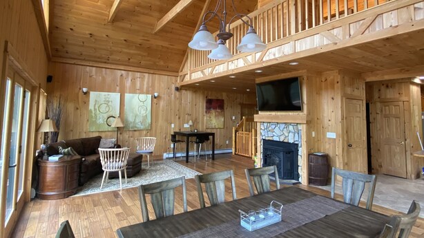 Un interior espacioso de madera con techo alto, un comedor con una mesa grande y sillas, una chimenea y un balcón con barandilla.