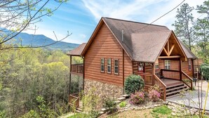A wooden cabin with a stone foundation, a porch, and a view of a forested mountain range.