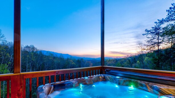 A hot tub with a wooden deck and glass walls, overlooking a forested landscape.