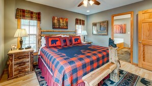 A bedroom with a large bed, a ceiling fan, a wooden nightstand, a lamp, a mirror, and a window with curtains.