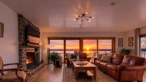 A living room with a fireplace, leather sofa, wooden coffee table, and large windows overlooking a sunset. - SPACIOUS and Stylish Lakefront Retreat, STUNNING Views, Firepit! (Fox Lake)
