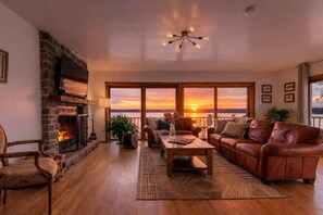 Una sala de estar con chimenea, sofá de cuero, mesa de centro de madera y grandes ventanas con vistas a un atardecer.