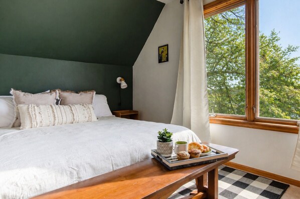 Un dormitorio con cama, una mesita de noche con lámpara, una ventana con vista a árboles y una bandeja con muffins y una taza.