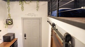 Huoneessa on kerrossänky, mikroaaltouuni puuhyllyllä, valkoinen ovi ja roikkuvia viherkasveja.