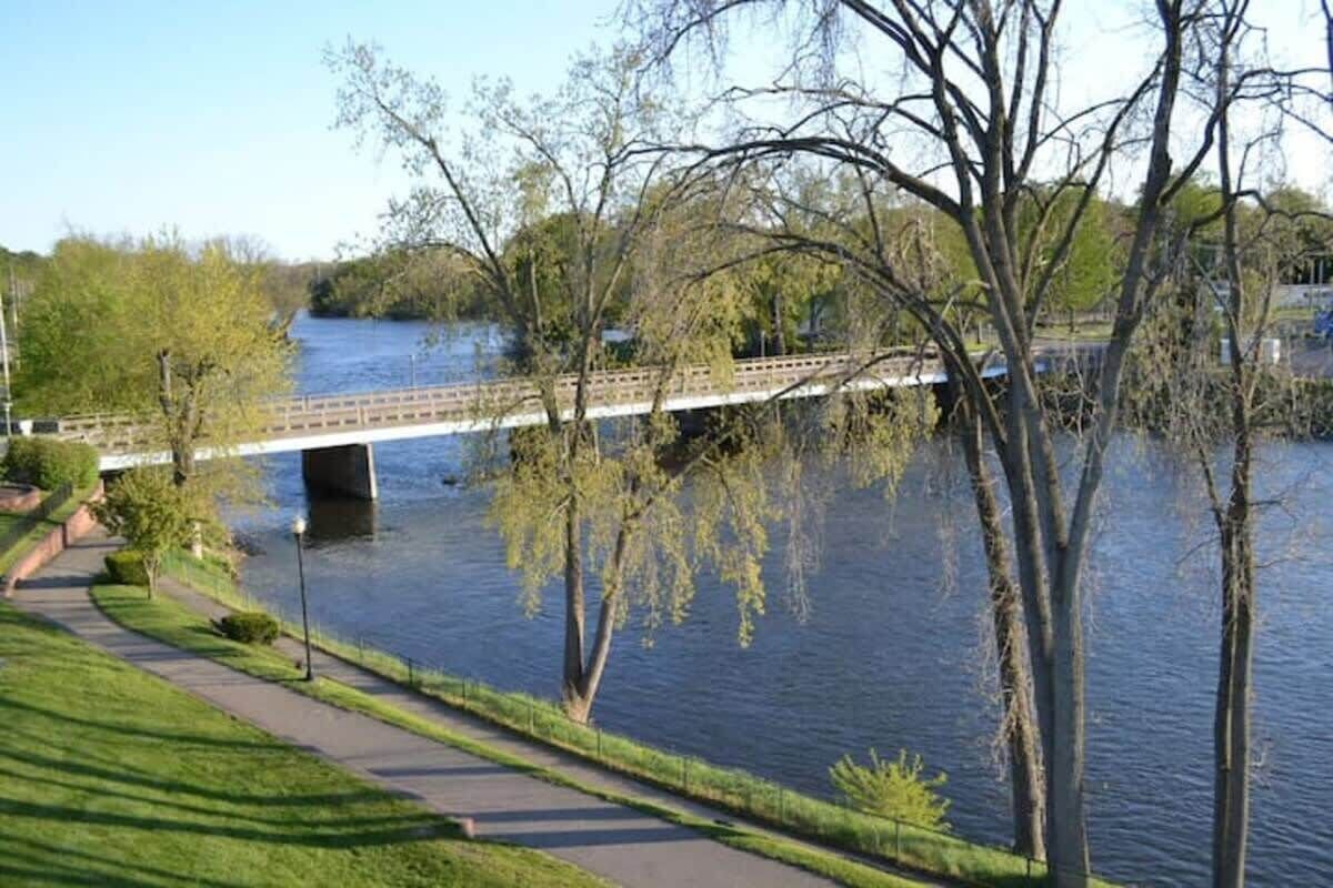 Une promenade au bord de la rivière, avec un pont, des arbres et un ciel dégagé.