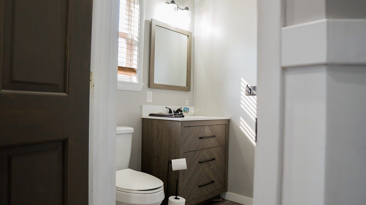 Une salle de bain comprenant des toilettes, un meuble en bois avec des tiroirs, un miroir et une fenêtre avec des stores.