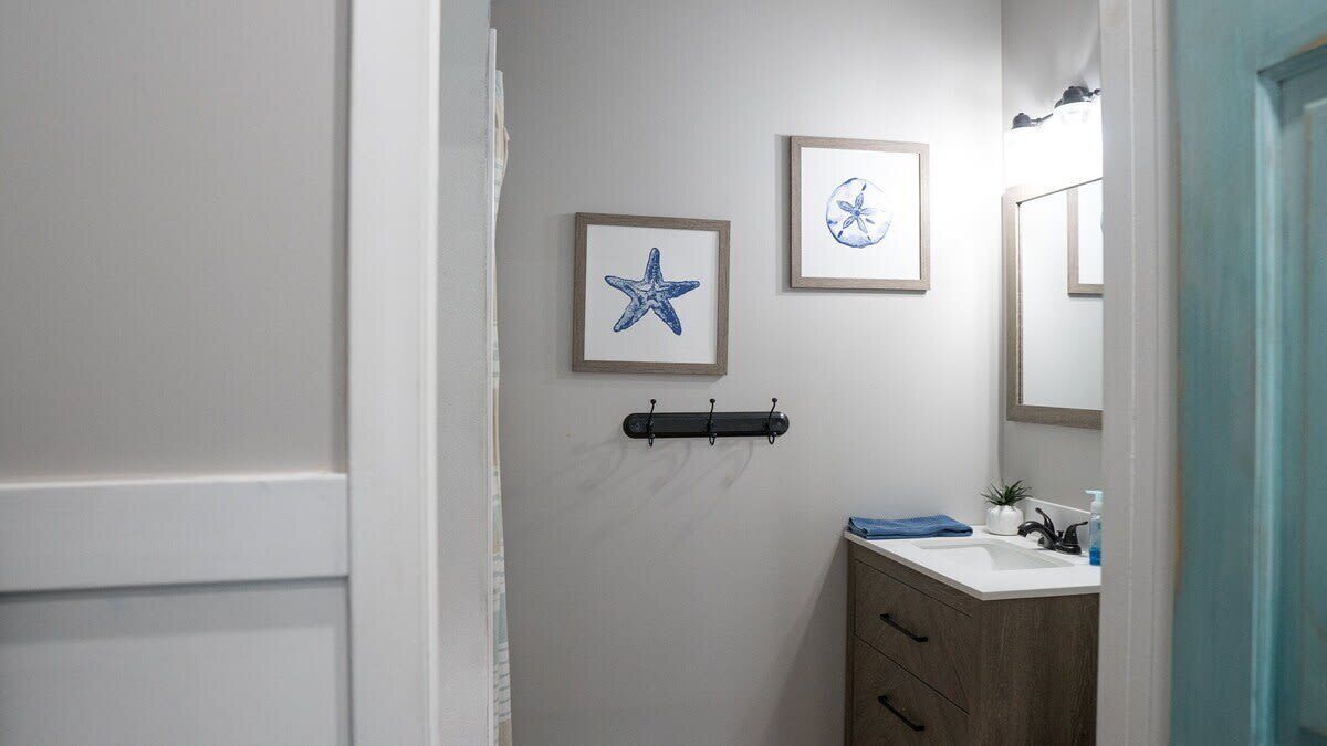 A bathroom with a white wall, a framed starfish picture, a framed flower picture, a black towel rack, a wooden cabinet, and a mirror.