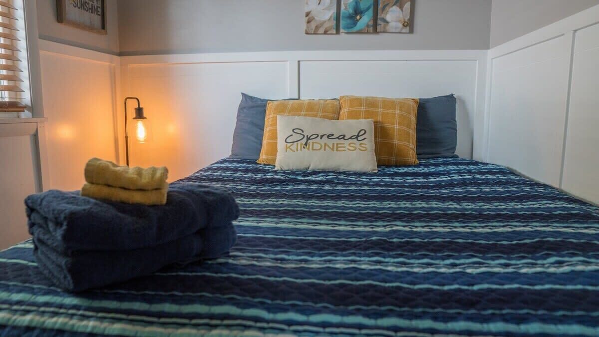 Un lit bien fait, recouvert d’une couvre-lit à motifs, un oreiller orné de l’inscription « Répandons la gentillesse », et une serviette pliée posée sur le côté gauche.