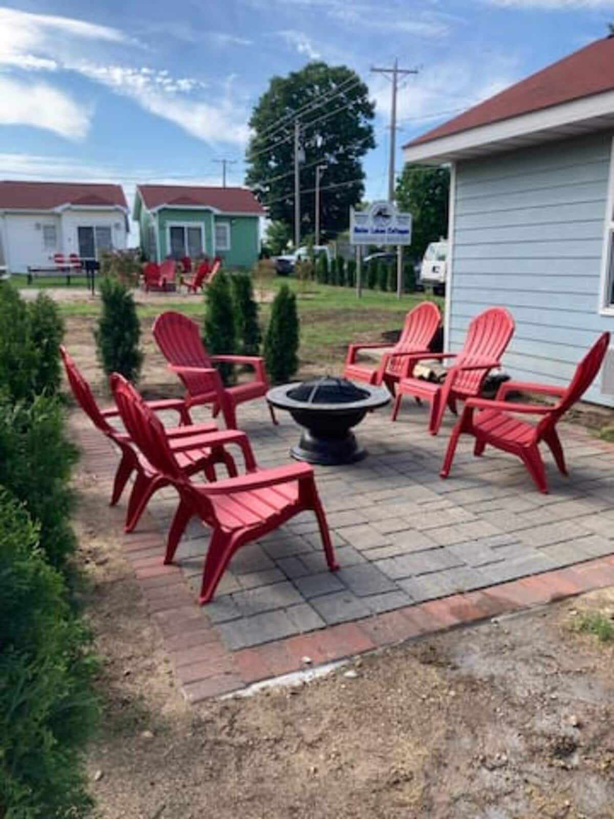 A patio area with red Adirondack chairs arranged around a fire pit, a brick pathway, and a small garden with potted plants.