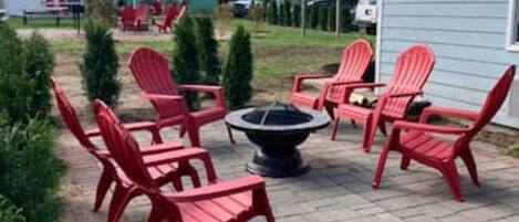A patio area with red Adirondack chairs arranged around a fire pit, a brick pathway, and a small garden with potted plants.
