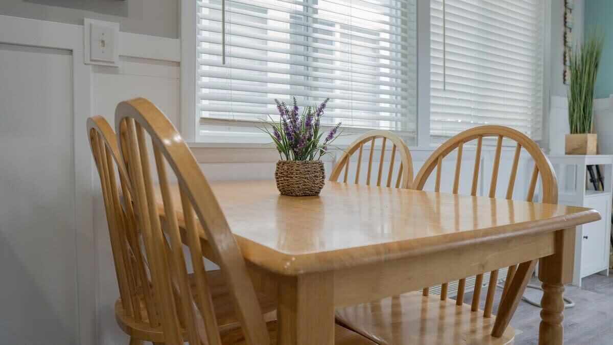 A wooden dining table with four chairs, a woven basket with purple flowers, and white blinds in the background.