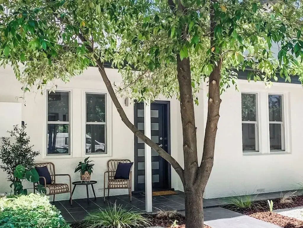 A white building with a tree in front, two wicker chairs, and a small table with a plant.