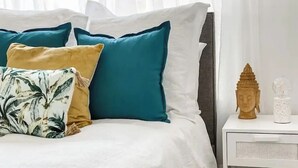A bed with teal and mustard pillows, a white bedspread, and a white nightstand with a Buddha statue and a glass container. - Stylish duplex in Prime Location! (Wagga Wagga)