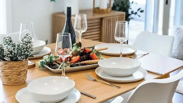A dining table set with white plates, bowls, and cutlery, accompanied by a bottle of wine and glasses. - Stylish duplex in Prime Location! (Wagga Wagga)