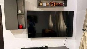 Ein modernes Wohnzimmer mit einem an der Wand befestigten Flachbildfernseher, einer dunkelgrauen Unterhaltungseinheit darunter und einem kleinen Regal mit Dekorationsobjekten.