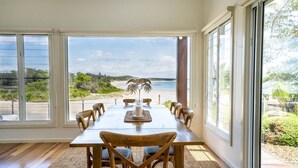 Un espace repas comprenant une table et des chaises, de grandes fenêtres offrant une vue sur la plage et l’océan, et un plancher en bois.