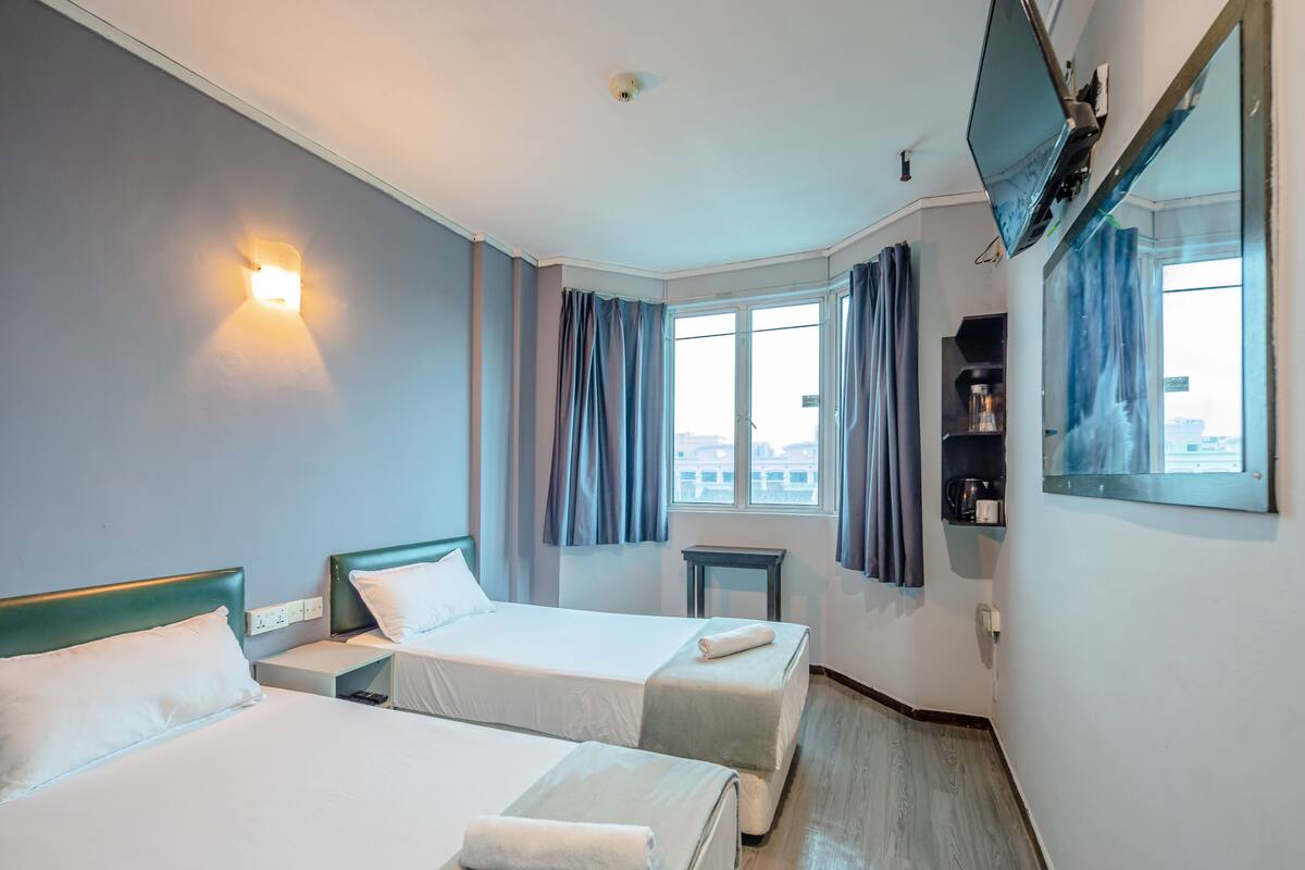 Ein Hotelzimmer mit zwei Betten, einem Fernseher, einem Fenster mit Vorhängen, einem kleinen Tisch und einem Regal mit Gegenständen.