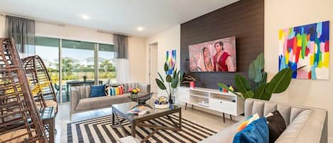 A modern living room with a sofa, coffee table, and TV. There is a balcony with a view of houses.