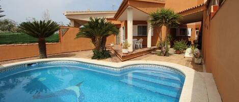 A swimming pool with a tiled edge, surrounded by a patio area with chairs and potted plants.