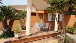 Uma área externa coberta com piso de azulejos, cadeiras de plástico branco e uma mesa.