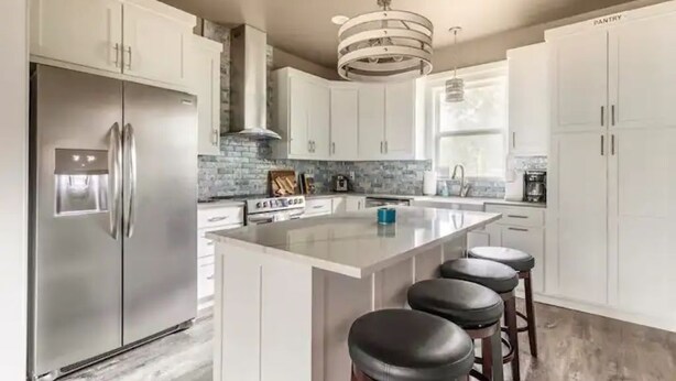 A modern kitchen with white cabinets, a stainless steel refrigerator, a central island with bar stools, and a hanging light fixture.