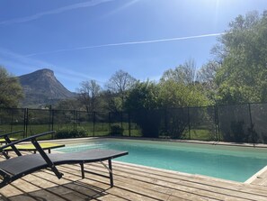 Une piscine dotée d’une terrasse en bois et de chaises longues, entourée d’une clôture et d’arbres, avec une montagne en arrière-plan.