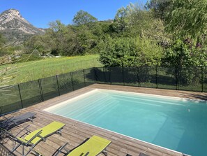 A rectangular swimming pool with a wooden deck, surrounded by a black mesh fence, and a mountain in the background.