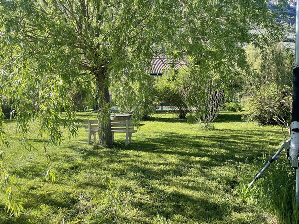 A grassy area with a bench and trees, a building with a tiled roof in the background, and a pole with a camera on the right.