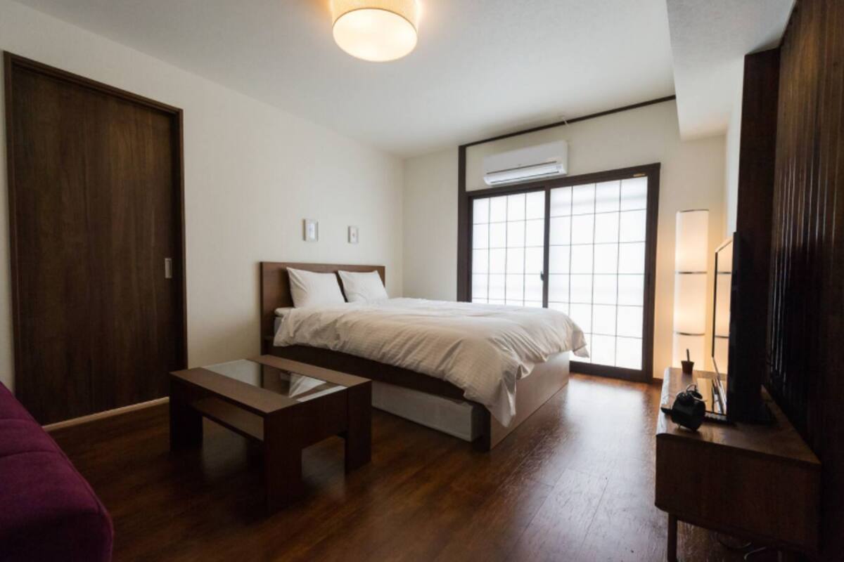 Quarto de hotel com uma cama, um banco de madeira, uma escrivaninha, uma televisão e uma janela grande com persianas.