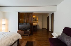 A modern hotel room with a wooden floor, a bed, a sofa, a TV, and a desk with a lamp.