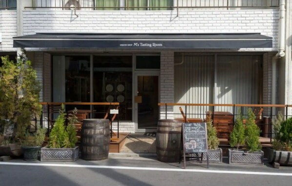 A brick building with a sign reading "M's Tasting Room" above the entrance. There are wooden barrels and potted plants outside.