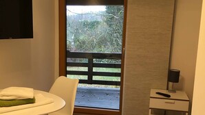 A room with a wooden floor, a white table with a green mat, a white chair, a small white nightstand with a lamp, and a sliding door leading to an outdoor balcony with a wooden railing. - Pension Jägerschenke (Luhe-Wildenau)