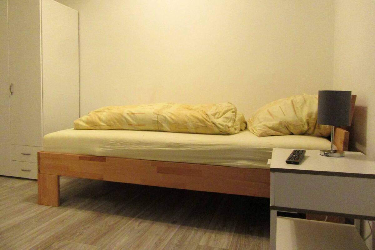 A small, single-bed room with a wooden frame, a bedside table with a lamp, and a wardrobe.