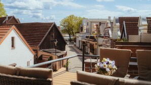 A terrace with wicker furniture and a flower arrangement, overlooking a street with traditional buildings and a clear sky. - Alex 12 "Meeresleuchten" 1.O.G.  (Rostock)