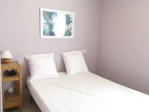 Uma cama de solteiro com roupa de cama branca, uma mesa de cabeceira com abajur e um quadro pendurado na parede.