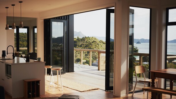 A modern kitchen with a large island, a dining area with a wooden table and chairs, and a balcony with a view of the sea and mountains. - A view like none other (Cooks Beach)