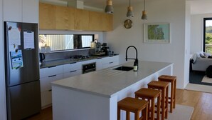 A modern kitchen with white cabinets, a large island with bar stools, stainless steel appliances, and pendant lighting. - A view like none other (Cooks Beach)