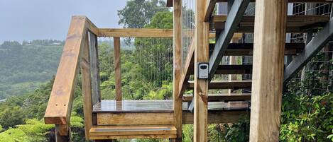 Una escalera de madera que conduce a un balcón con baranda de metal, rodeado de vegetación exuberante y vistas a árboles distantes.