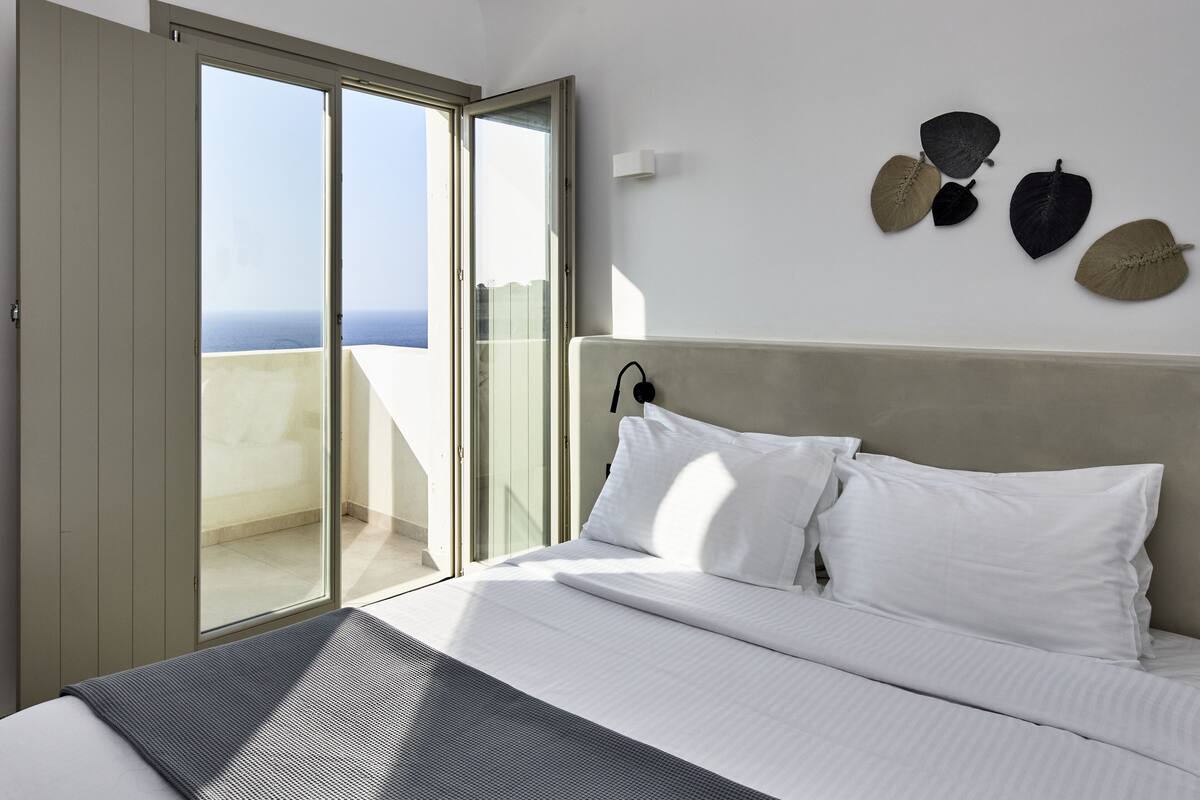 Hotelkamer met een bed, witte kussens, uitzicht door een schuifdeur en wanddecoraties van bladeren.
