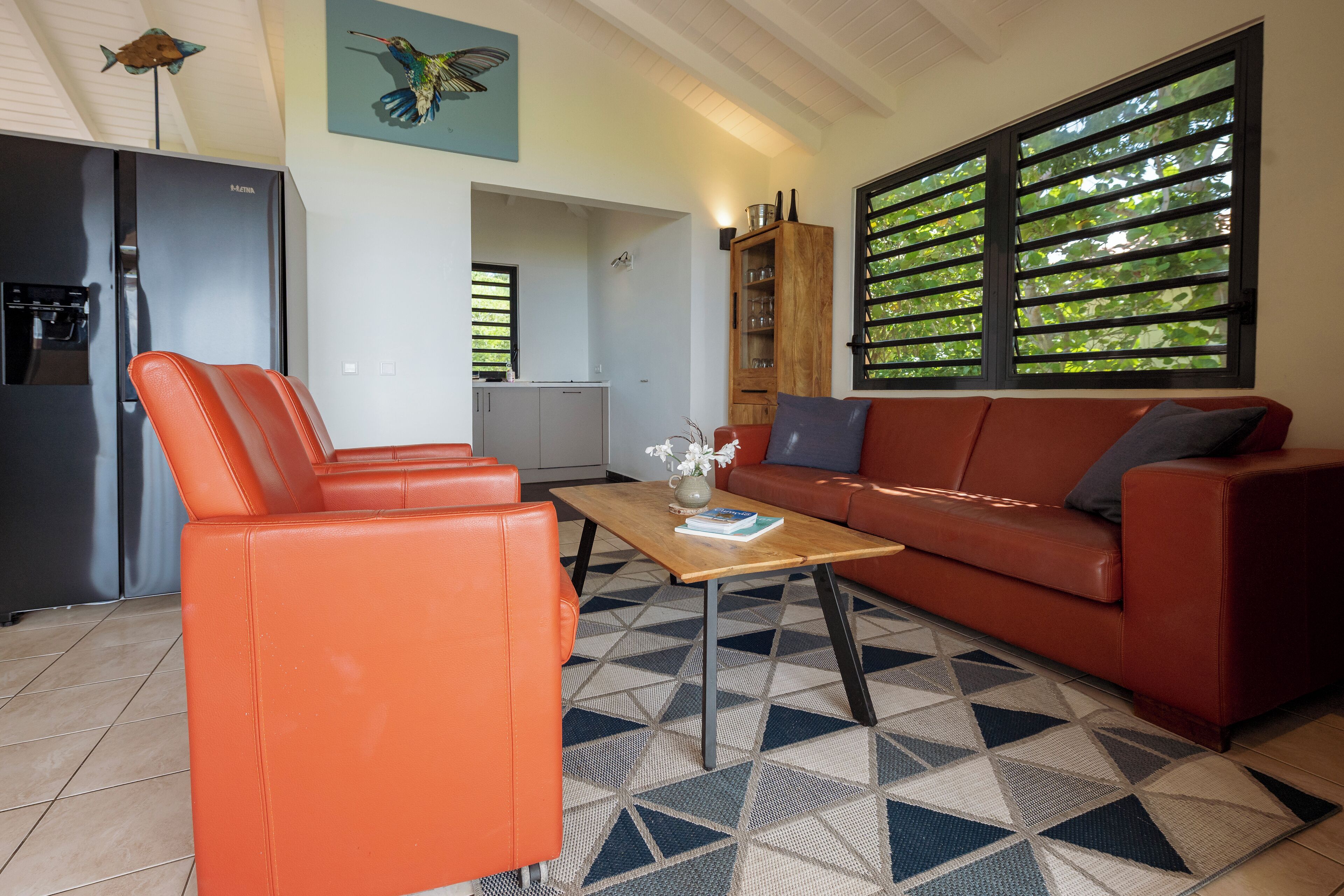 Un salon moderne avec un canapé, une table basse et un fauteuil rouge. On y trouve également un coin cuisine avec un réfrigérateur et une fenêtre équipée de stores.