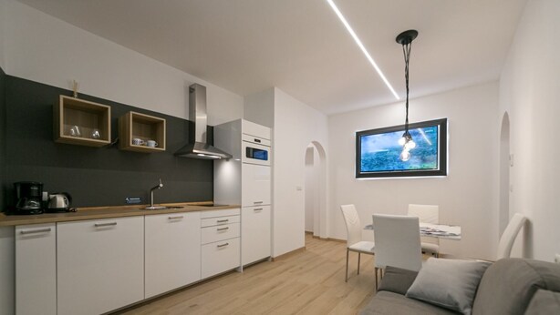 Una cucina moderna con mobili bianchi, un piano di lavoro in legno e un forno integrato. C'è una zona pranzo con tavolo e sedie, e un divano grigio nel soggiorno.
