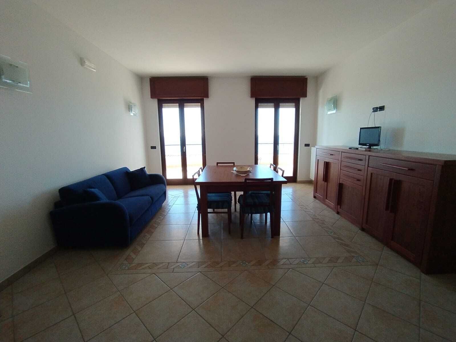Ein Wohnzimmer mit blauem Sofa, einem Esstisch mit Stühlen, einem Fernseher auf einer Kommode und einer Küchenzeile mit Schränken.
