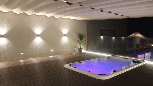 A hot tub on a wooden deck with a glass railing, surrounded by walls with built-in lighting. - Hotel Rajhans Abhinandan (Bhopal)
