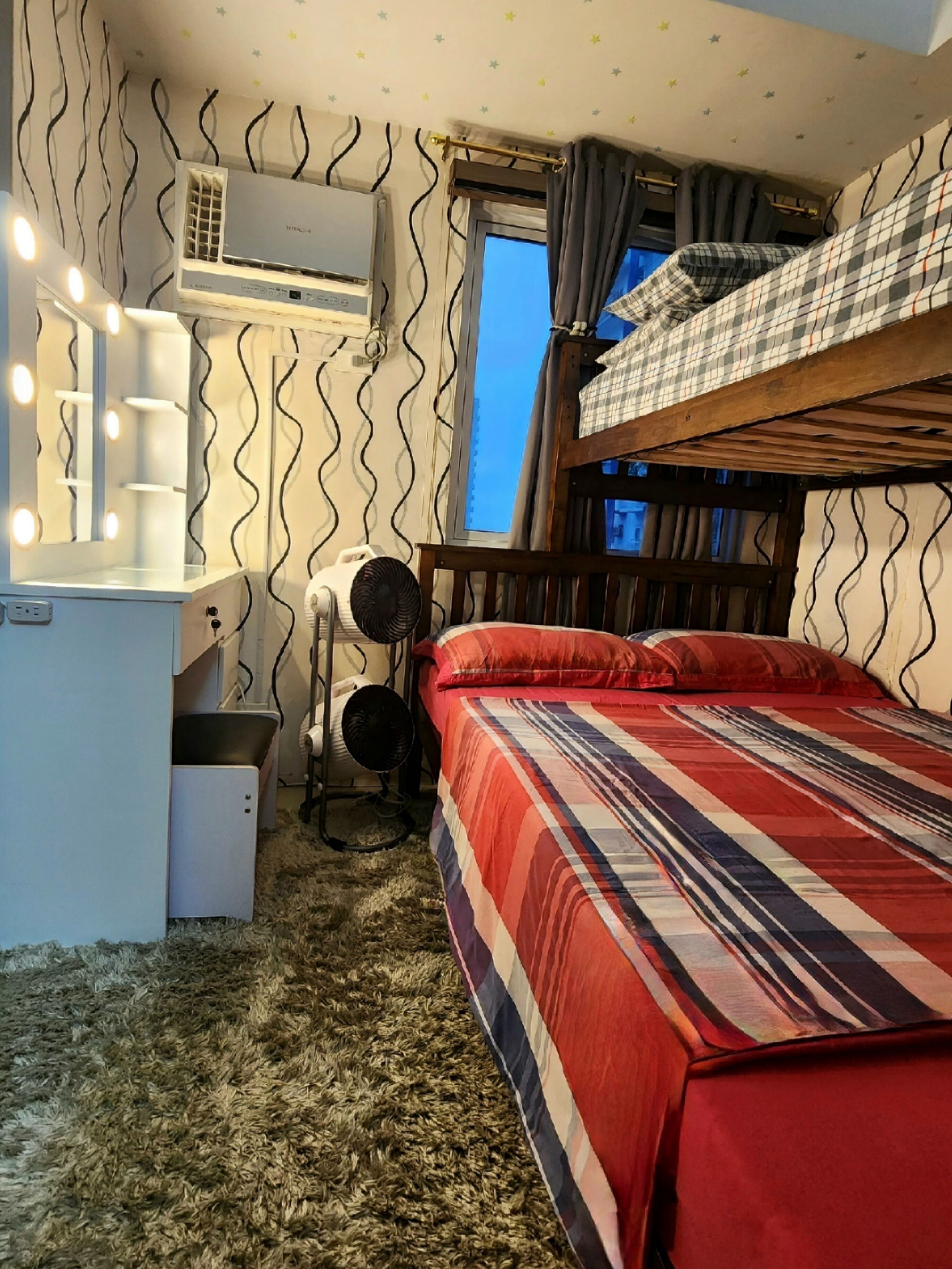 Een compacte slaapkamer met een stapelbed, een bureau met spiegel en een ventilator.