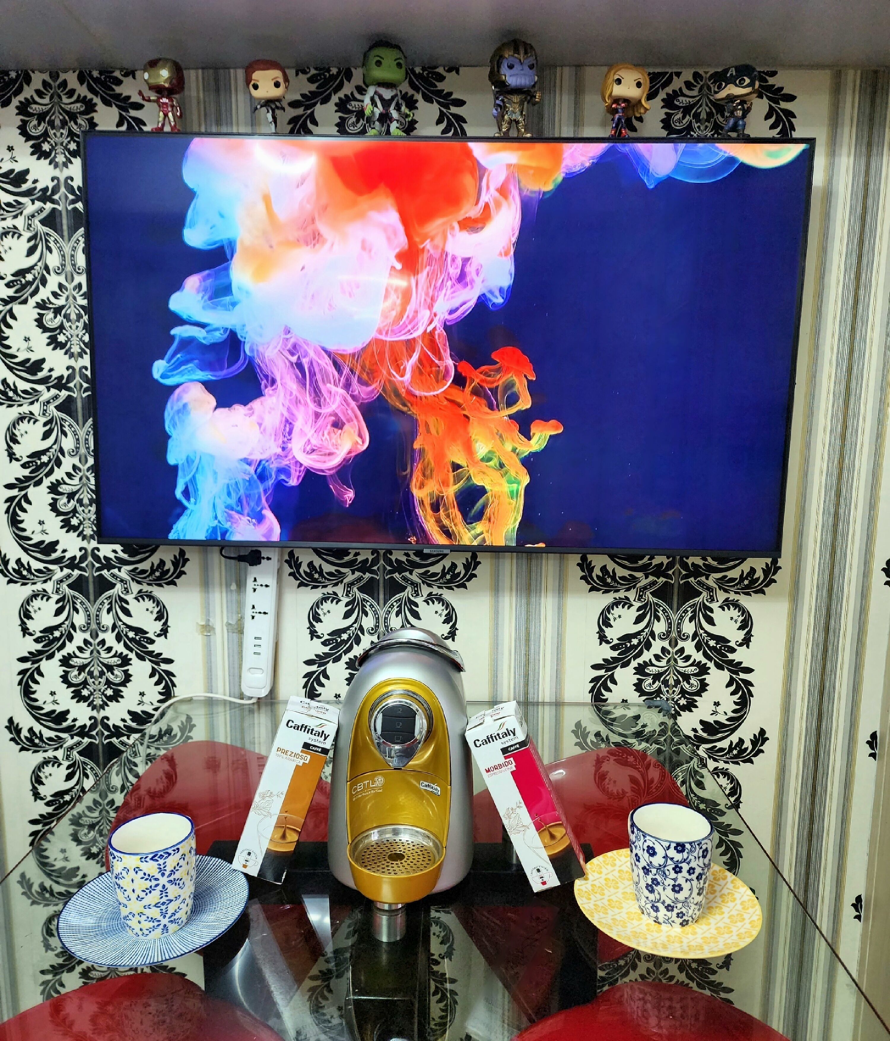 Een kamer met een flatscreen televisie waarop kleurrijke abstracte kunst wordt getoond, een koffiezetapparaat en twee kopjes op een tafel.