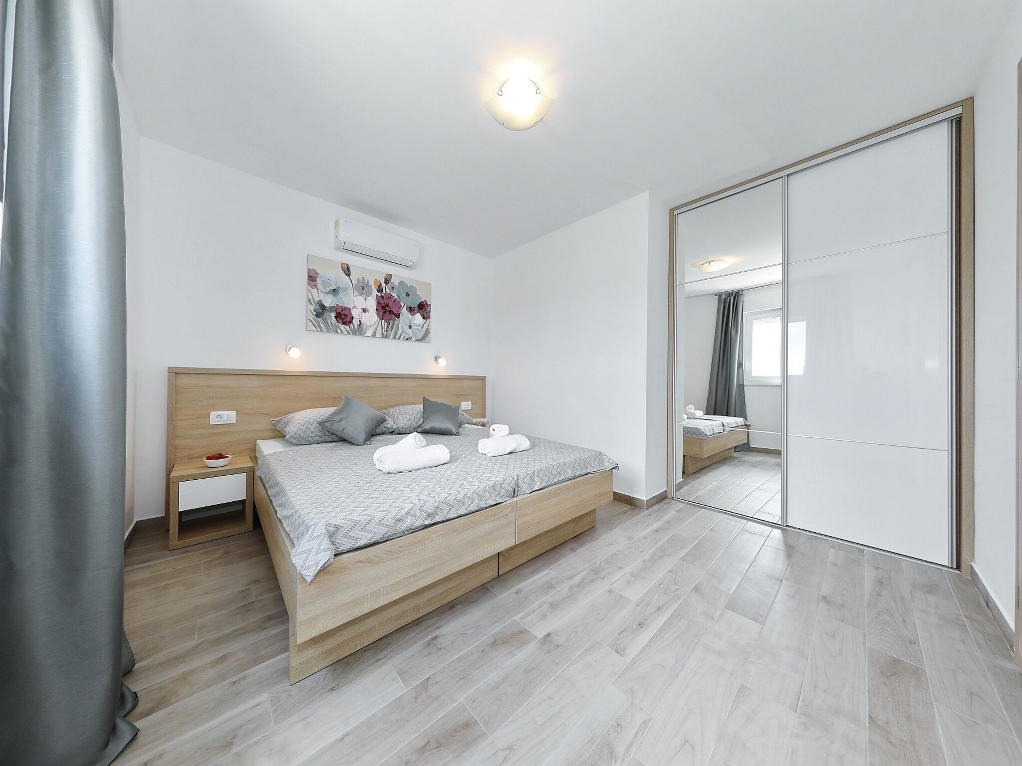A modern bedroom with a wooden bed, a nightstand, a wall-mounted air conditioner, and a large mirror.