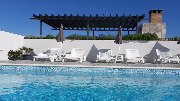 Una piscina trasparente con lettini e ombrelloni, un pergolato e una struttura in pietra sullo sfondo.